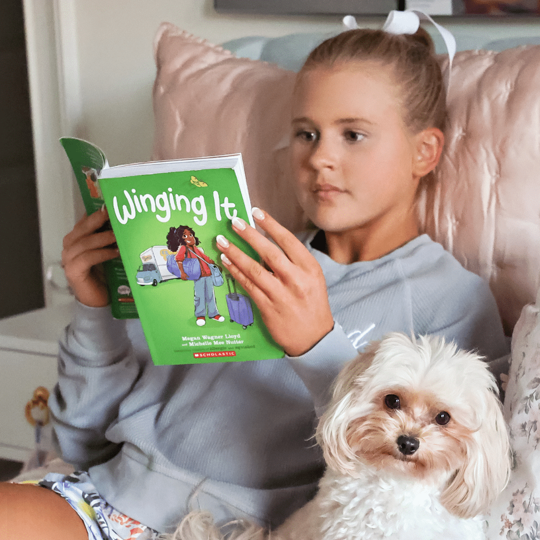 A young girl sits on a bed reading Winging It by Melissa Wheeler and Lisa Siberry. She wears a light grey hoodie, and a small white dog with fluffy fur sits beside her, beside a notebook titled New You Checklist.