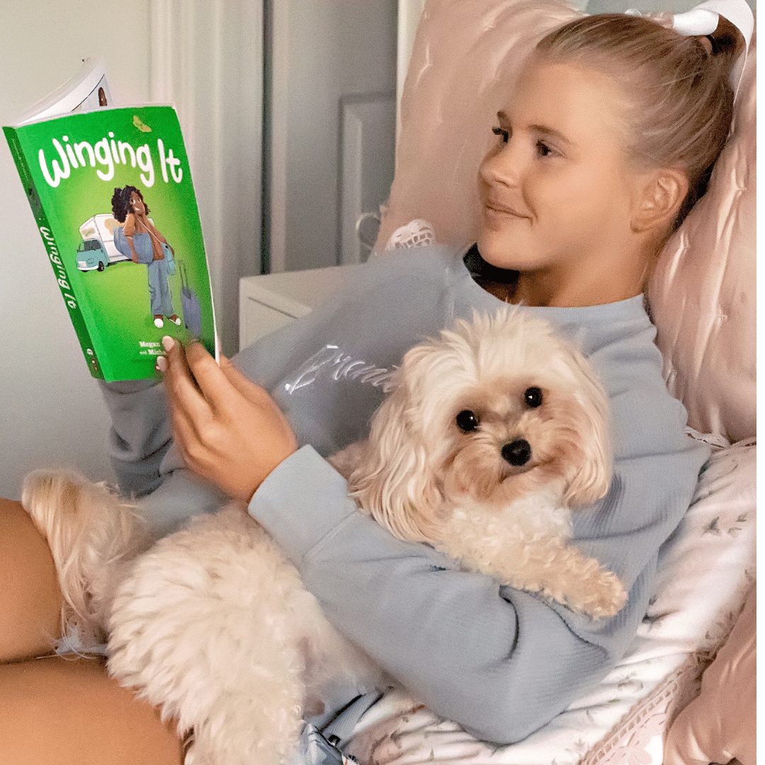 A girl with blonde hair, wearing a light blue sweatshirt, sits on a bed and smiles while holding a fluffy white dog in her lap and reading her green book, Winging It—her New You Checklist for a cozy afternoon.