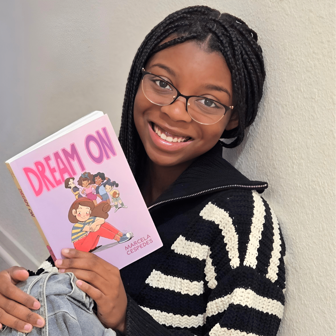 A smiling girl with glasses and braided hair holds a book titled Dream On by Marcela Cespedes, featuring illustrated children on the cover. She wears a black and white striped sweater and sits against a light-colored wall.