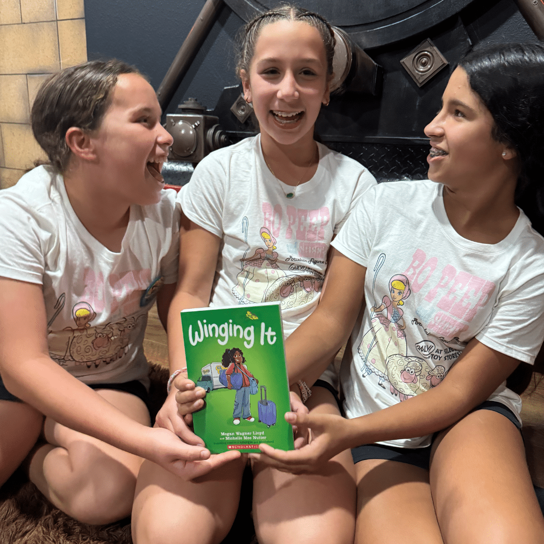 Three smiling girls sit together in matching T-shirts, holding up a green book titled Winging It with an illustrated girl on the cover. They look happy and engaged, ready to dive into this fall reset guide together.