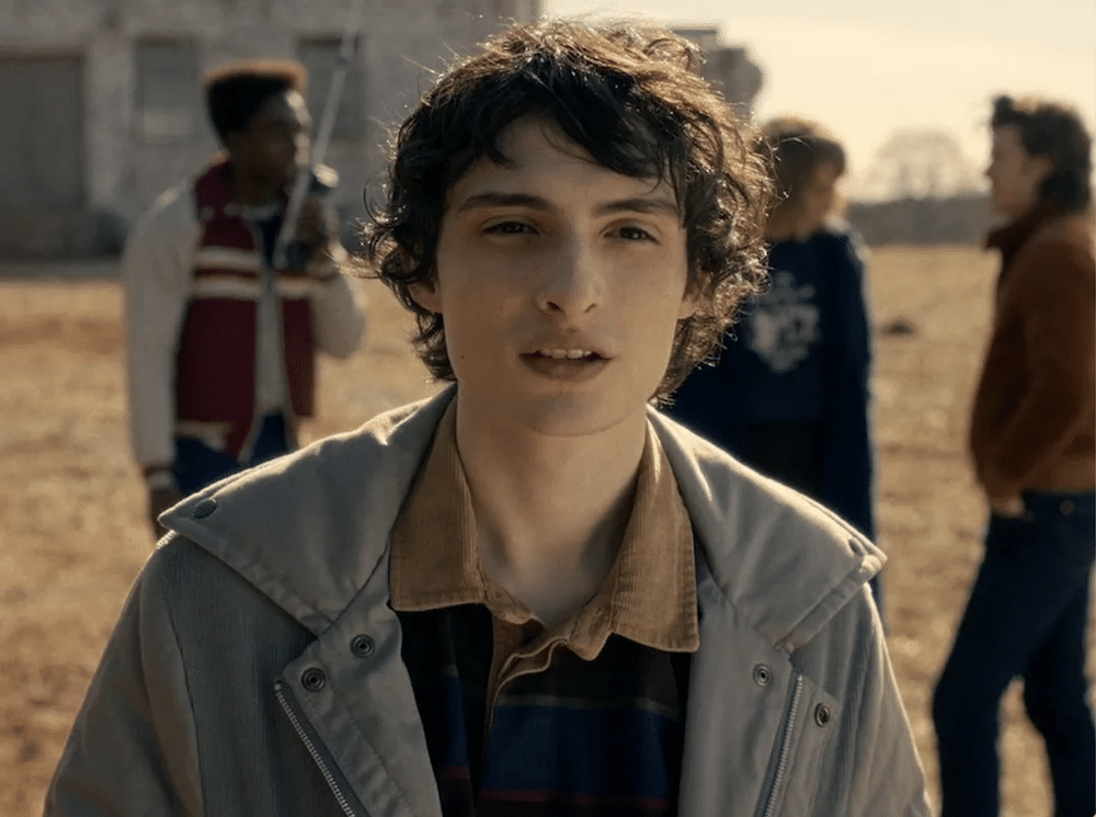 A teenage boy with wavy brown hair stands outdoors, looking at the camera like a Stranger Things character. Three other teens are blurred in the background, talking in a field with an old building behind them.