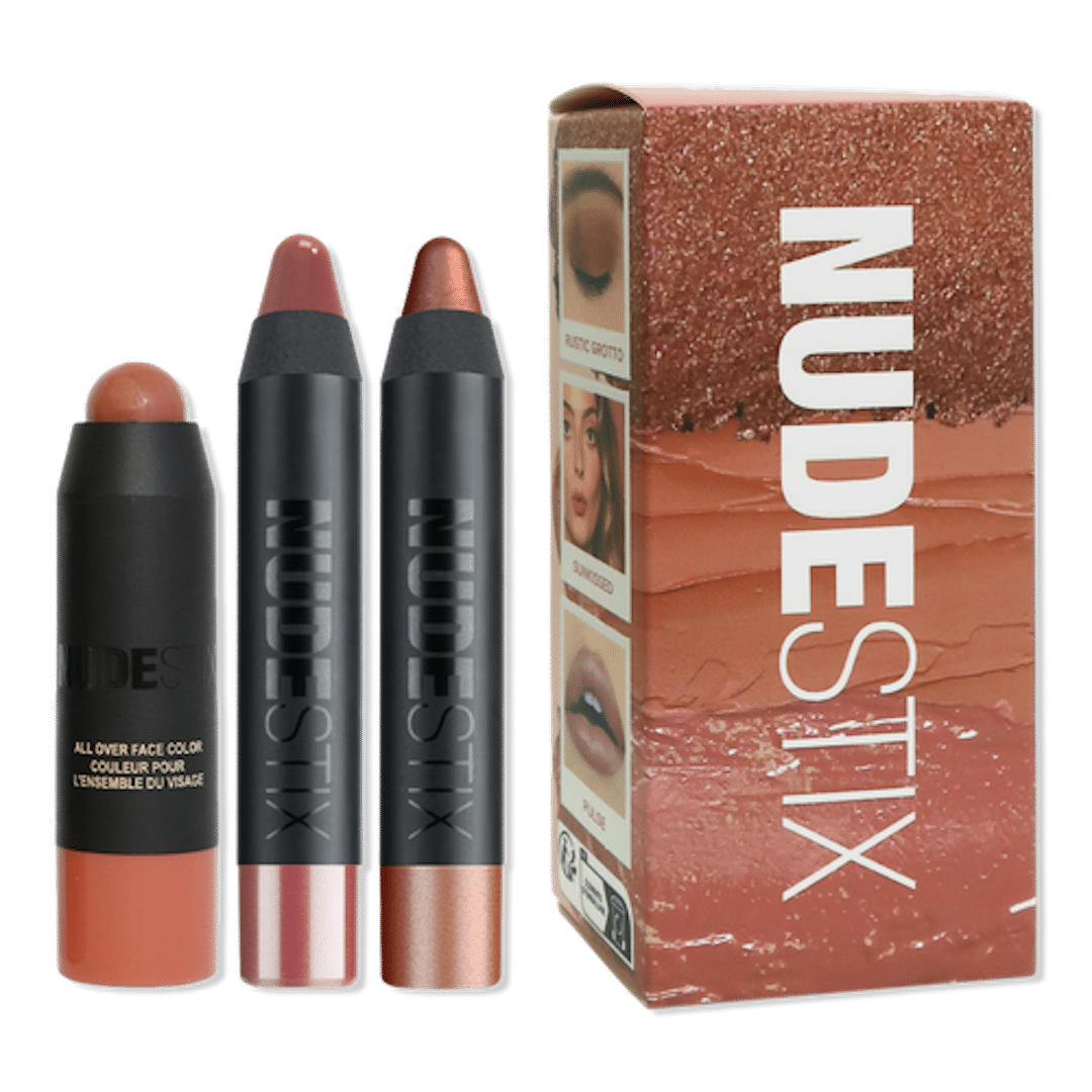 Three NUDESTIX makeup sticks—one in a rounded tube, two in pencil form—are displayed next to a NUDESTIX branded box with a shimmery nude design and swatches, serving up full-glam inspo worthy of The Life of a Showgirl.