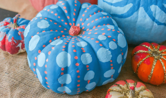 A blue pumpkin decorated with light blue dots and orange accents sits on burlap among other painted and glittered pumpkins in various colors, offering charming autumn home decor inspiration.