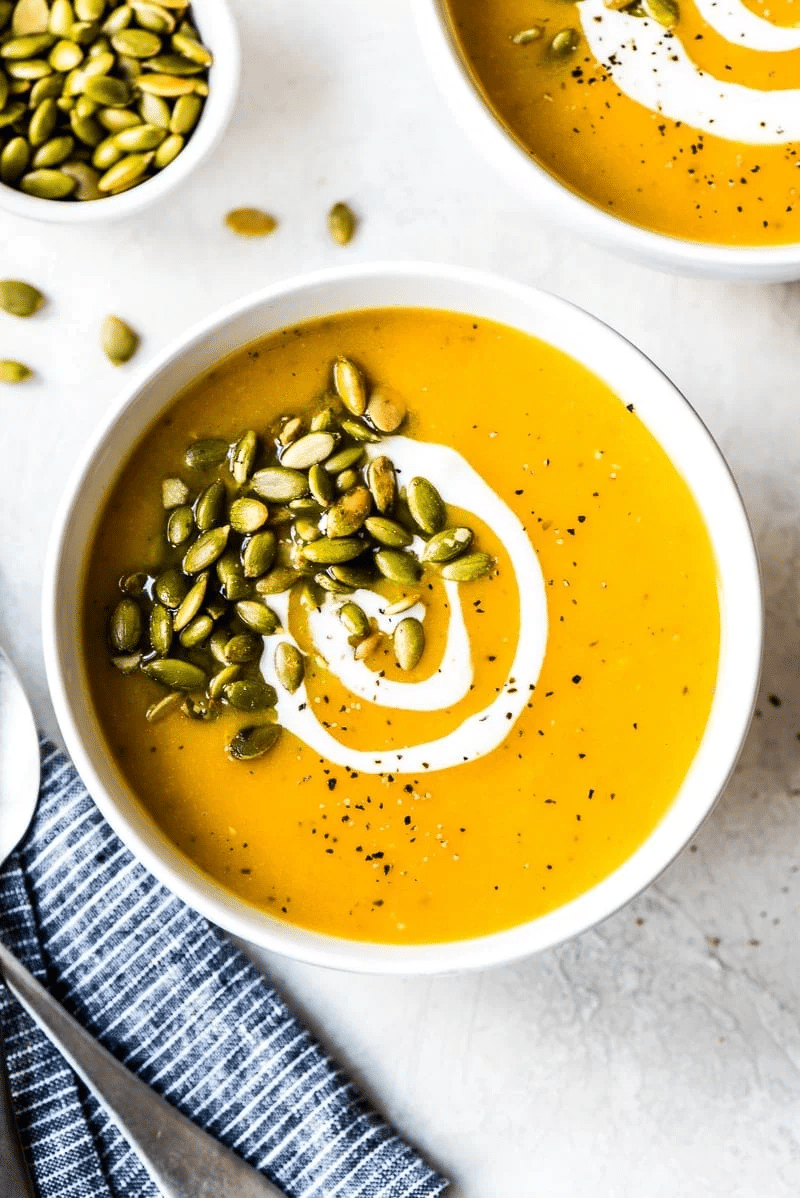 A white bowl of creamy orange soup topped with a swirl of cream, roasted pumpkin seeds, and black pepper, next to a striped napkin and a spoon. Another bowl and a small dish of seeds are partially visible.