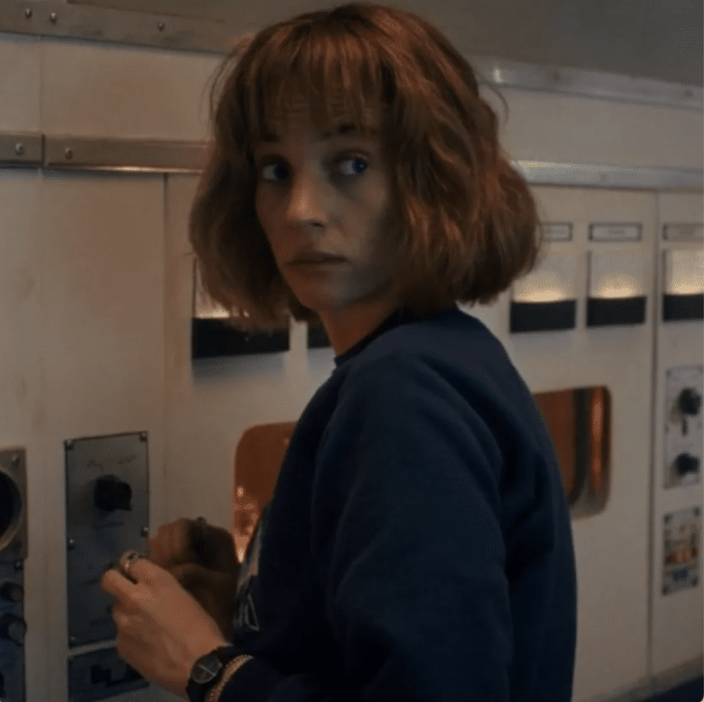 A character with short, wavy brown hair and bangs stands in front of control panels, turning to look over her shoulder with a concerned expression. She wears a dark blue sweater and a wristwatch, evoking the suspenseful vibe of Stranger Things.