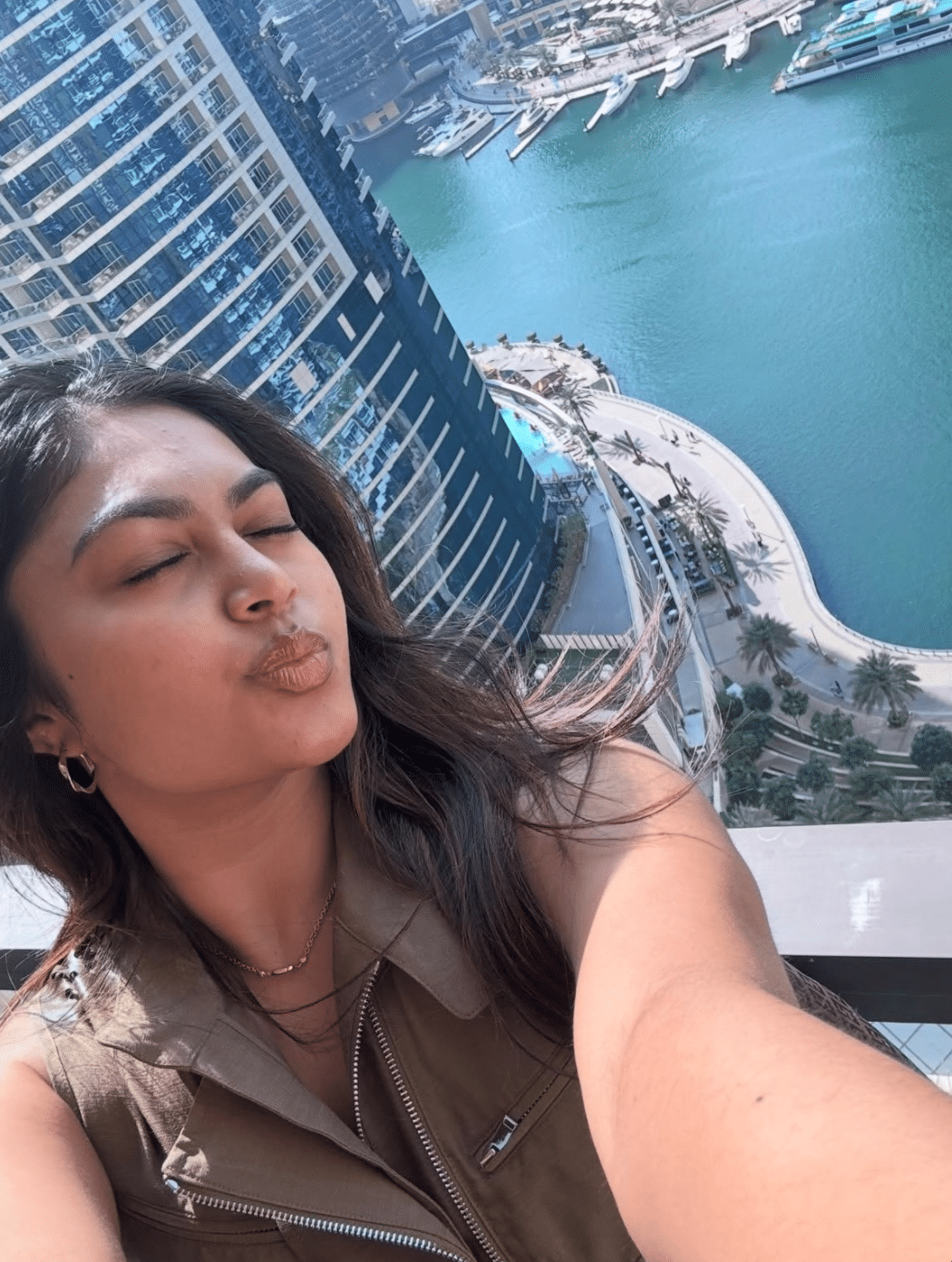 A woman with long dark hair takes a selfie while making a kissy face on a balcony, reminding us to stop comparing your love life to others', as she enjoys the view of tall buildings, a pool, palm trees, and a marina with boats below.