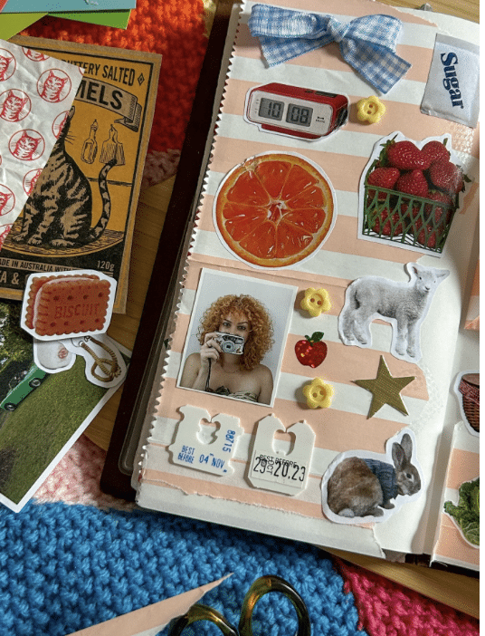 A scrapbook page decorated with stickers of fruit, animals, a thermometer, a camera, a bow, buttons, tickets, a barcode, and various colorful images, alongside yarn, scissors, and craft supplies.