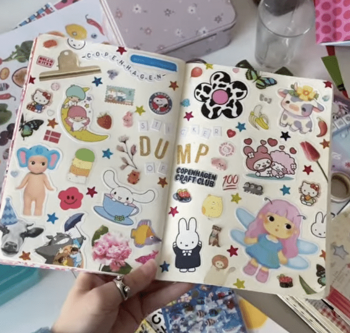 An open journal with colorful, playful stickers including cartoon animals, fruits, stars, bunnies, flowers, and text like Sticker Dump. A hand holds the journal above a cluttered desk.