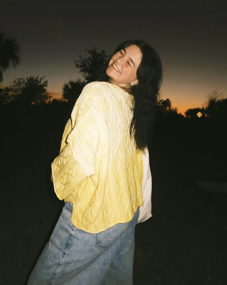 A woman with long dark hair, wearing a yellow sweater and blue jeans, stands outdoors at sunset, smiling and looking over her shoulder. The sky is dark with an orange glow on the horizon.