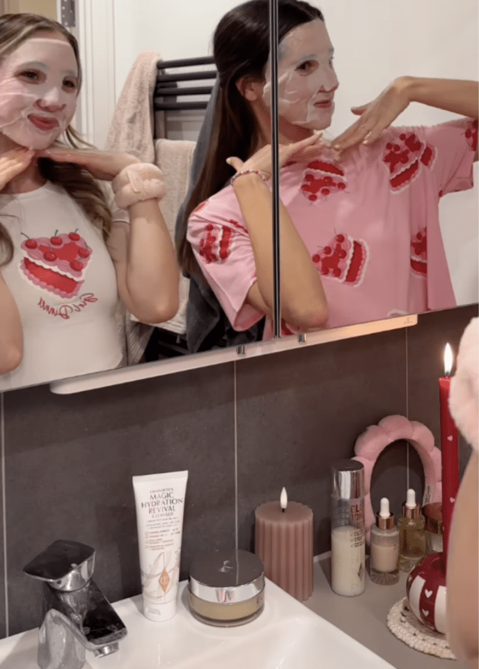 Two women in cake-print shirts and sheet face masks pose with hands under their chins by a bathroom mirror, surrounded by skincare products and candles—a perfect Galentine’s Day self-care night.