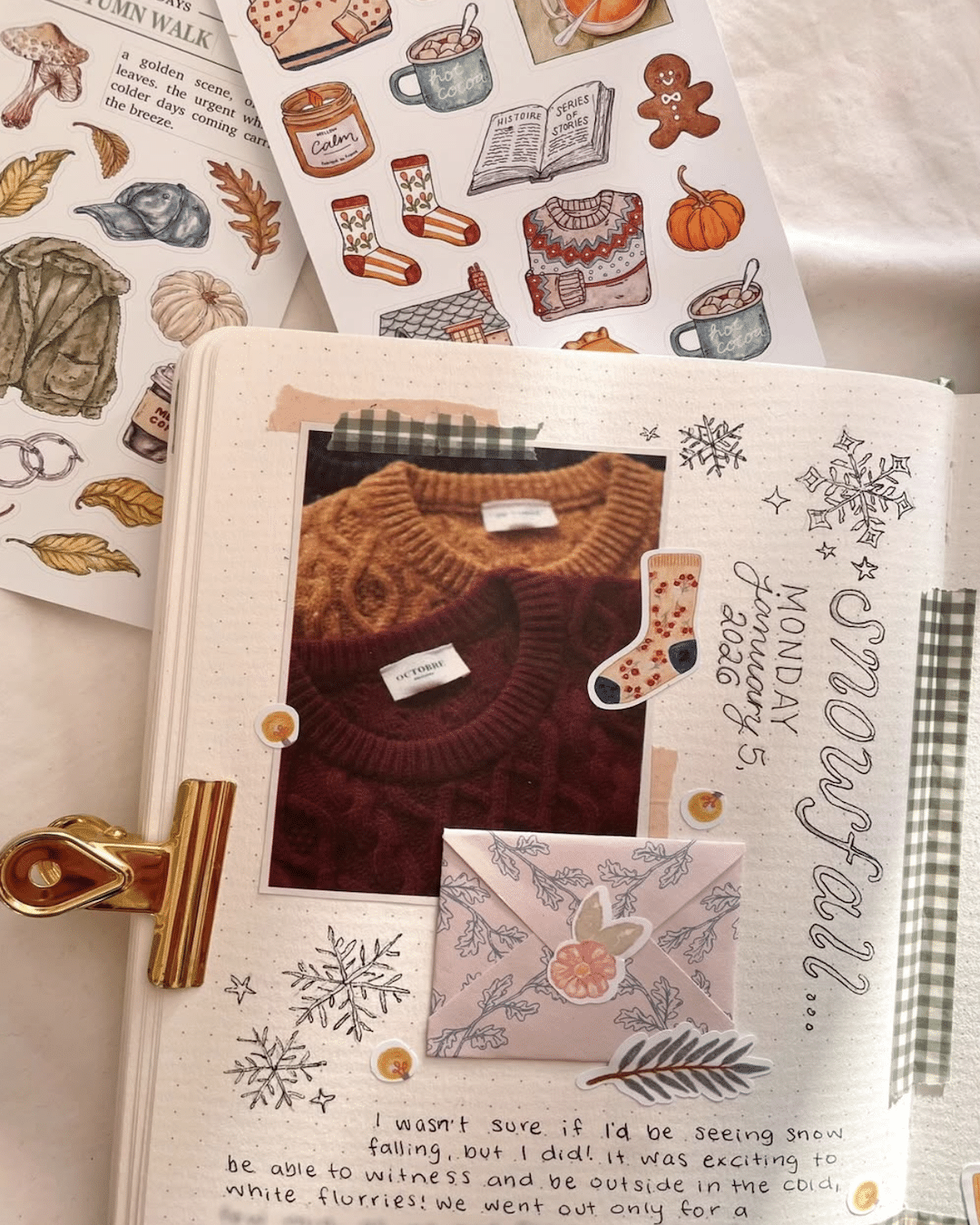 A scrapbook page decorated with stickers of sweaters, leaves, and mugs. There are photos of knit sweaters, handwritten notes about snowfall, and a pink envelope—all clipped together with a gold binder clip to inspire your New Year's journaling habit.