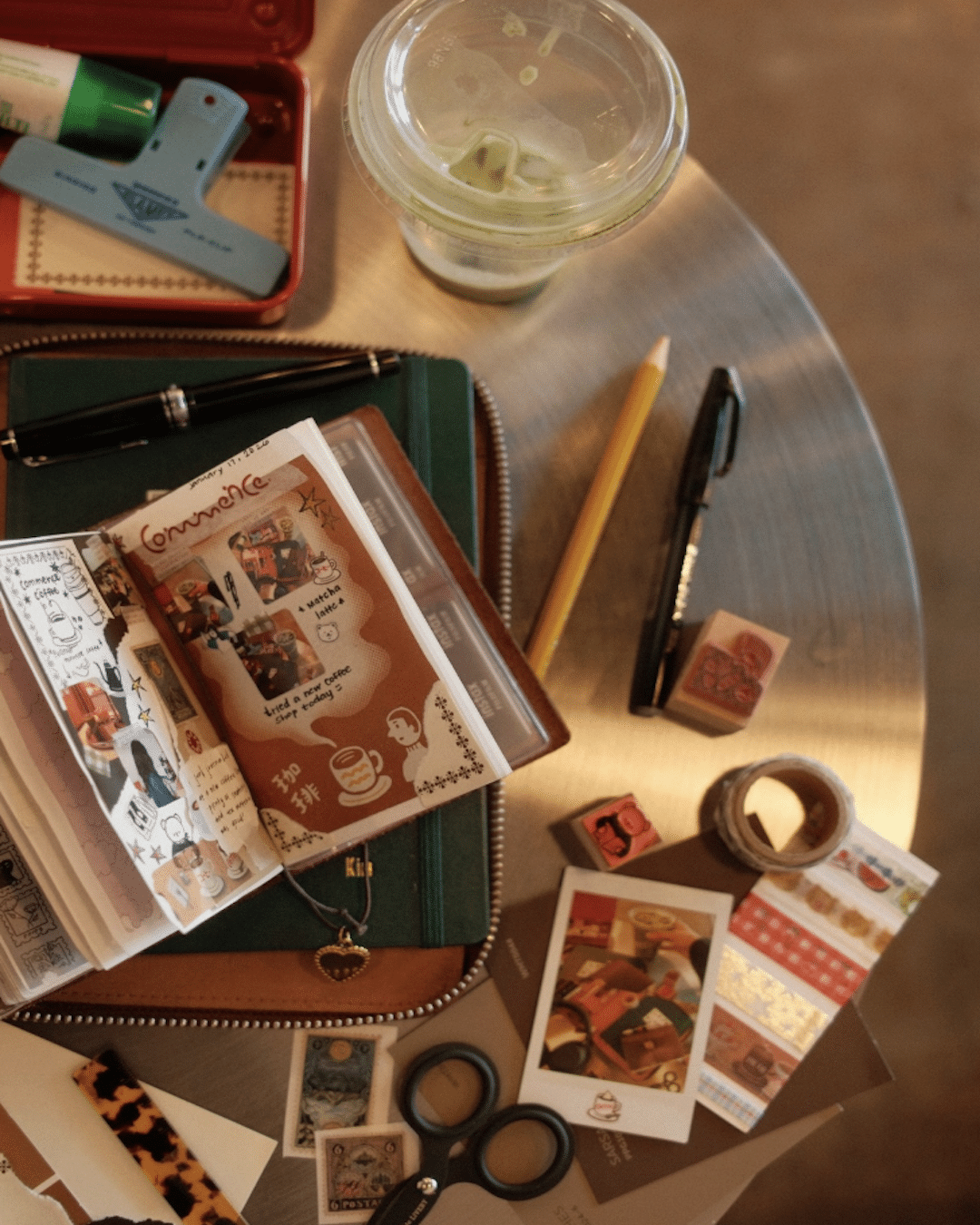 A round table with stationery items, perfect for New Year's journaling, featuring a journal open to decorated pages, pens, scissors, stickers, a rubber stamp, washi tape, and a green drink—just like the setups of lifestyle YouTubers.