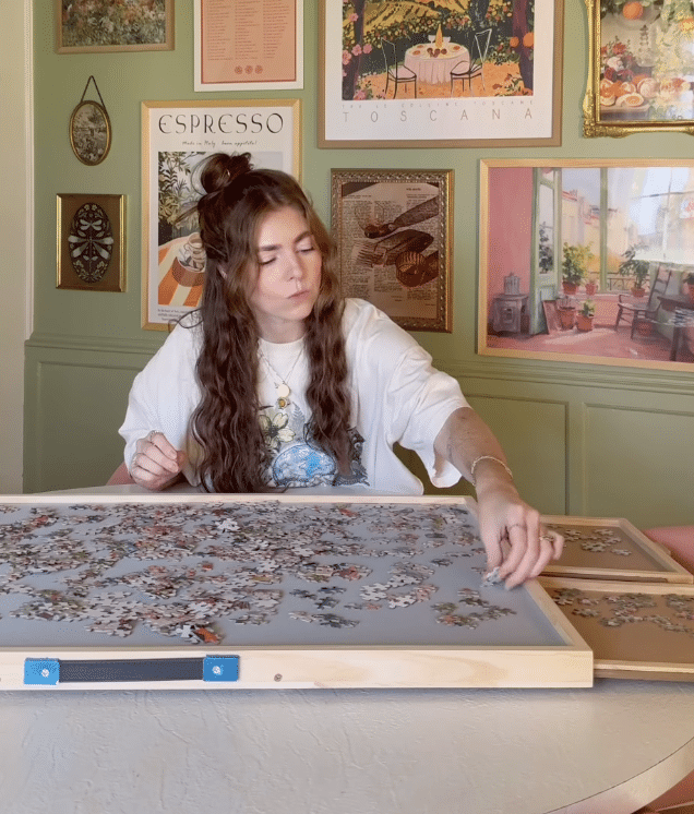 A woman with long brown hair sits at a table, assembling a jigsaw puzzle. She wears a white T-shirt with a graphic design. The background features colorful framed artwork and posters on green and beige walls.
