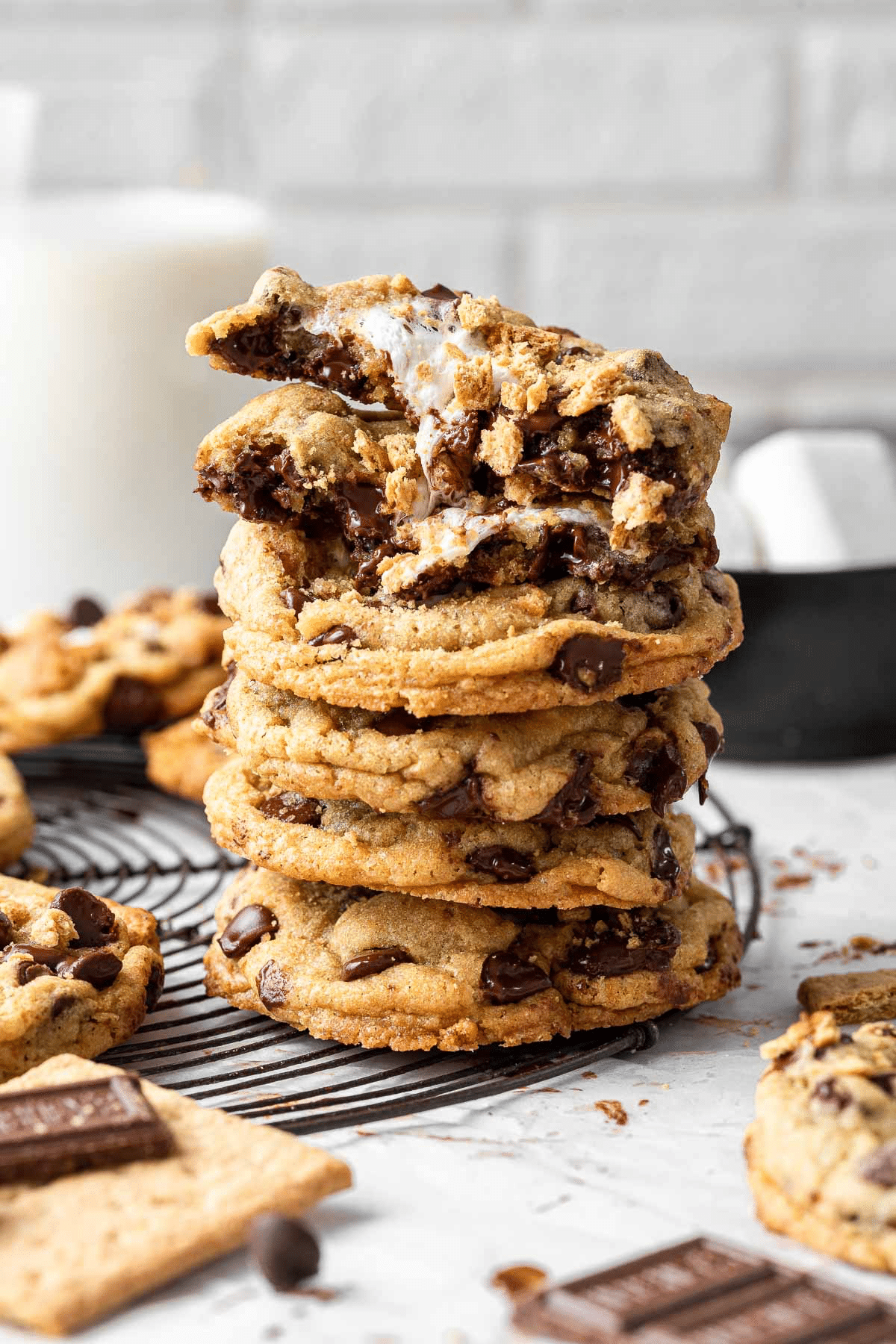 A stack of gooey chocolate chip cookies with melted marshmallow pieces, perfect for holiday baking, sits with the top cookie broken in half. More cookies and chocolate pieces are scattered on a cooling rack and surface nearby.