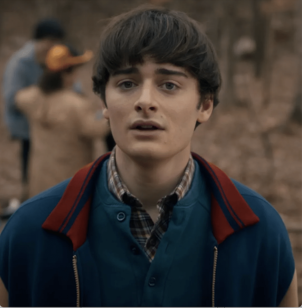 A young person with short brown hair, wearing a blue jacket with a red collar over a plaid shirt, stands outdoors in a wooded area with blurred people in the background.