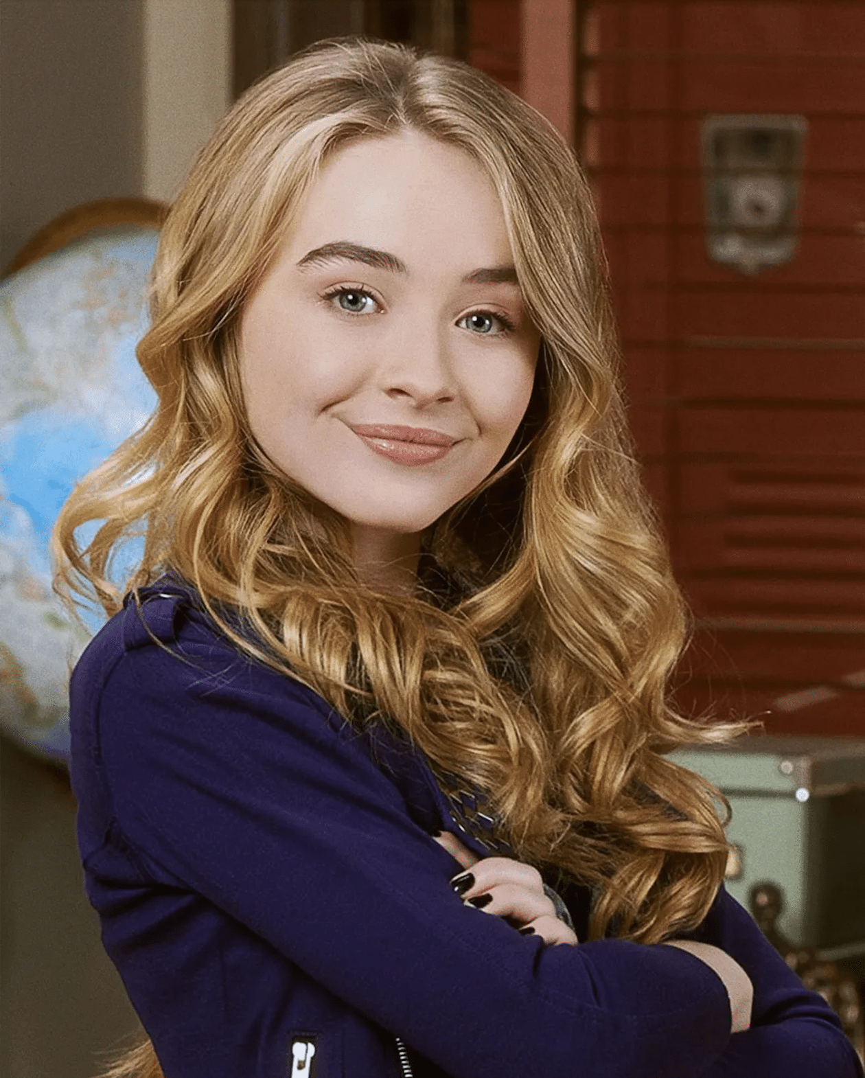 A young woman with long blonde wavy hair, wearing a navy blue jacket, stands with arms crossed and smiles confidently—showing How It Started and How It's Going. A globe and red blinds are visible in the background.